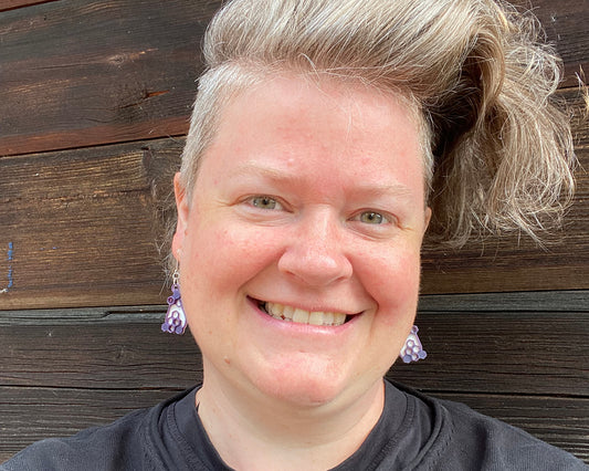A smiling model is wearing a pair of small white and purple earrings with a honeycomb design. 