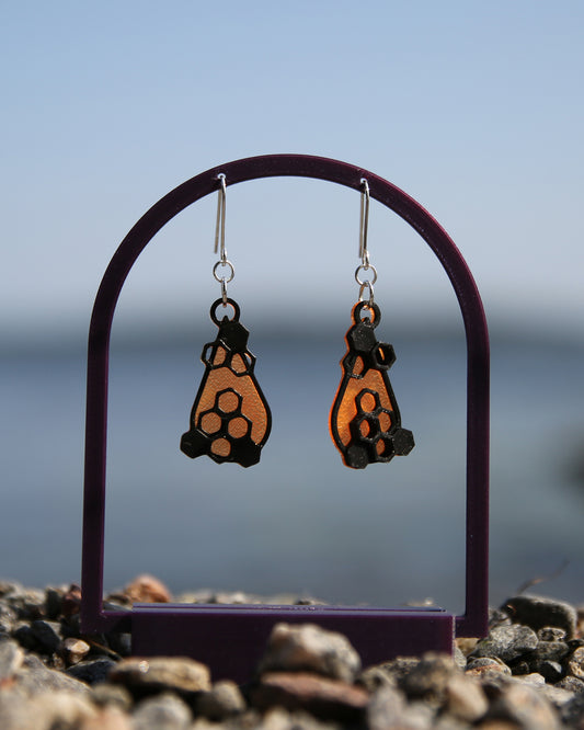 A pair of orange and black earrings with a honeycomb design, haning on a purple stand with a blurred natural background.