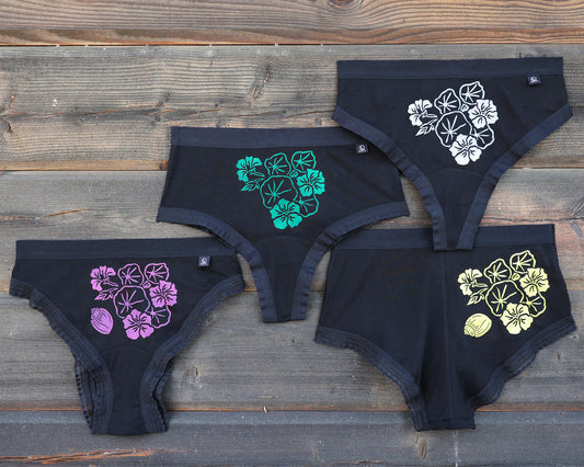 Four pairs of black underwear on a rustic wooden background, each featuring a different colored indian cress flower; magenta, green, white and yellow. This is the Indian Cress print! The magenta and yellow coloured version alos has a small printed shell.