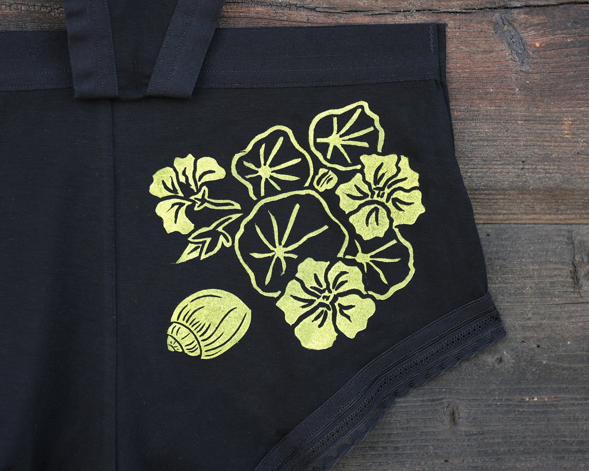 Close up of black underwear featuring a yellow graphic of indian cress flowers and leafs on the back, laid flat on a rustic wooden surface. There is also a yellow smaill shell.
