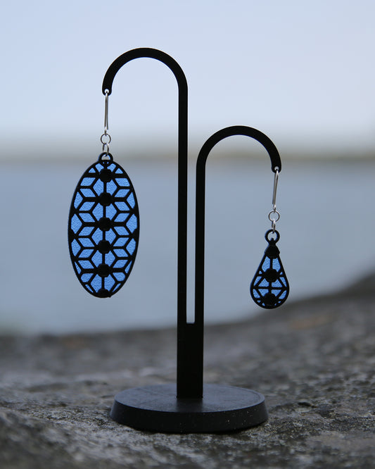 Two earrigs with a lace inspiered design, displayed on a black stand with a blurred natural background. The earring to the left is the original big size and the earring to the right is a small verion of the original design. They are both blue and black. 