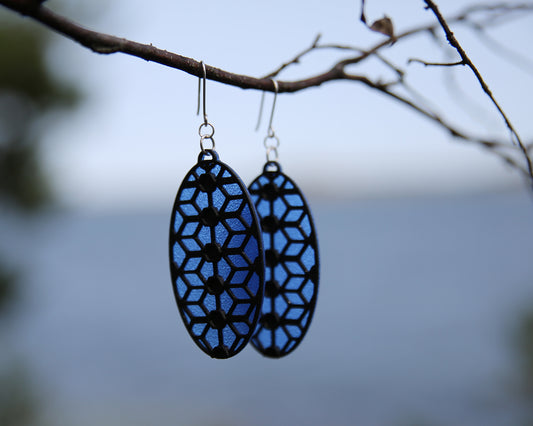 A pair of oval shaped earrings with a lace inspired design, hanging on a branch. The earrings have a blue translucent coloured background, and the pattern has a sparkling black colour.