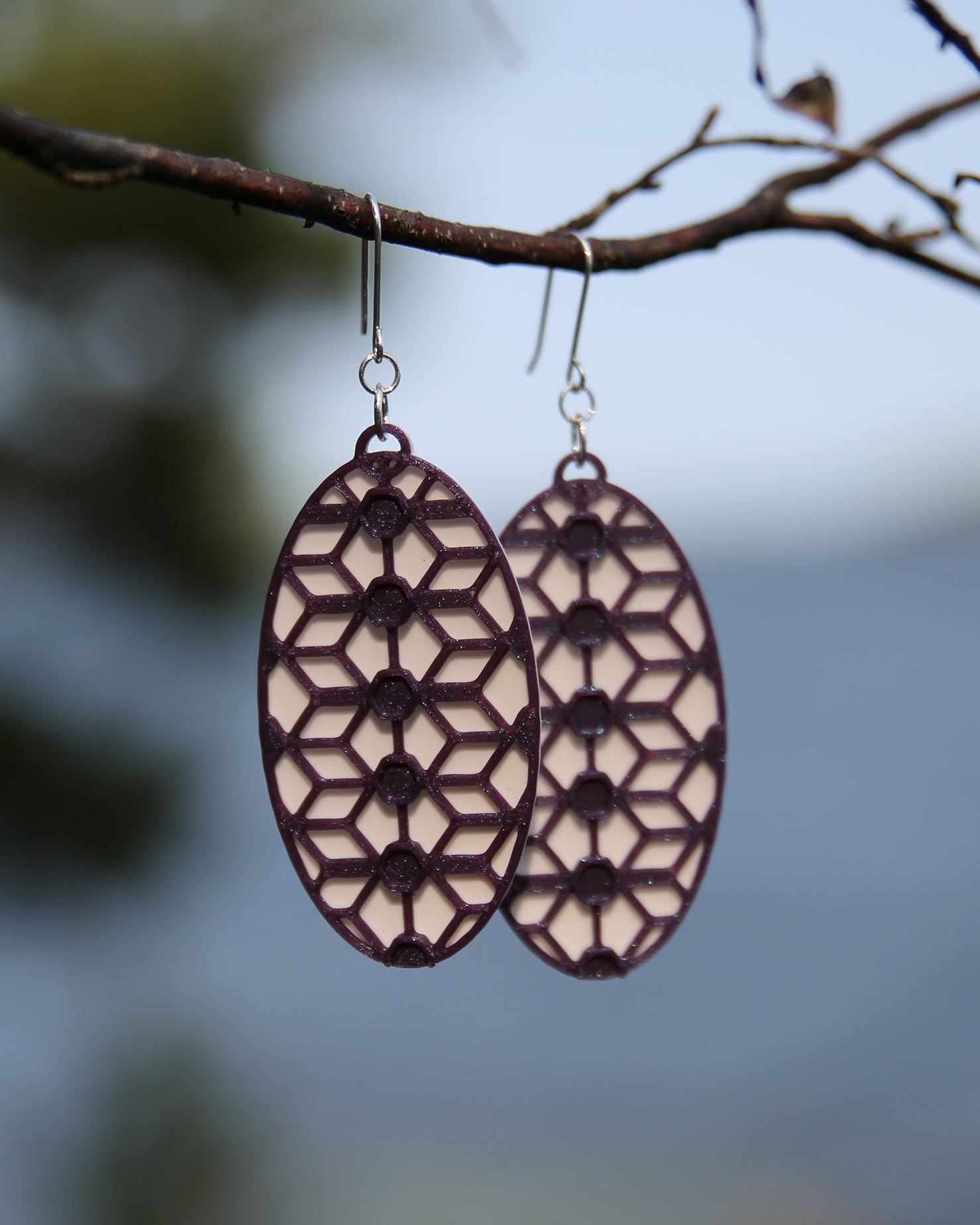 A pair of oval shaped earrings with a lace inspired design, hanging on a branch. The earrings have a white coloured background, and the pattern has a glittering purple colour.