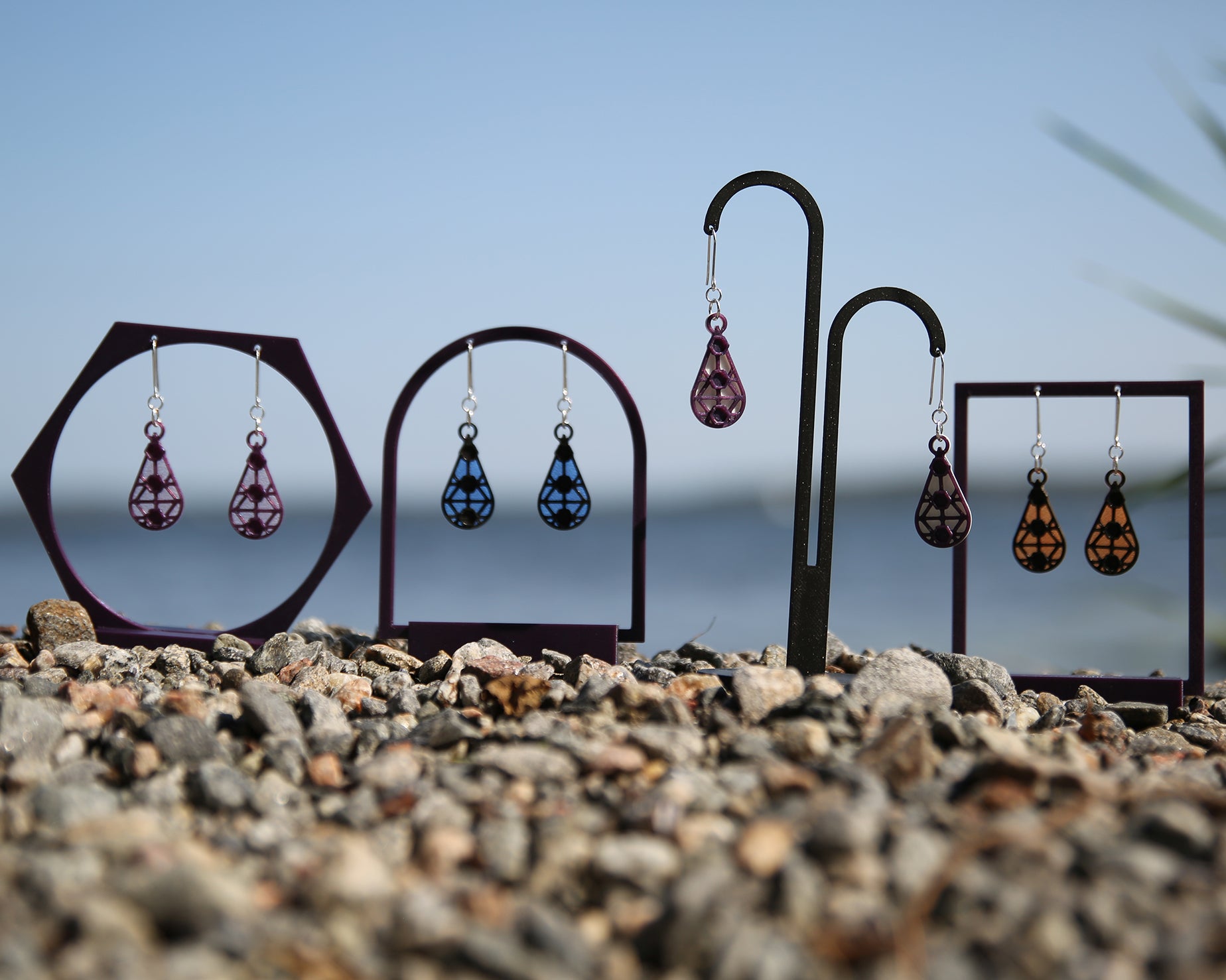 Colorful earrings with a lace inspiered design, displayed on stands against a blue sky and rocky ground.