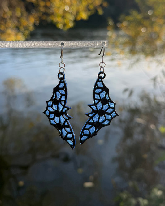 A pair of dangling earrings with a bat-shaped silhouette and a blue & black stained-glass like pattern, hanging from a clear stand against a blurred outdoor background of water and foliage.