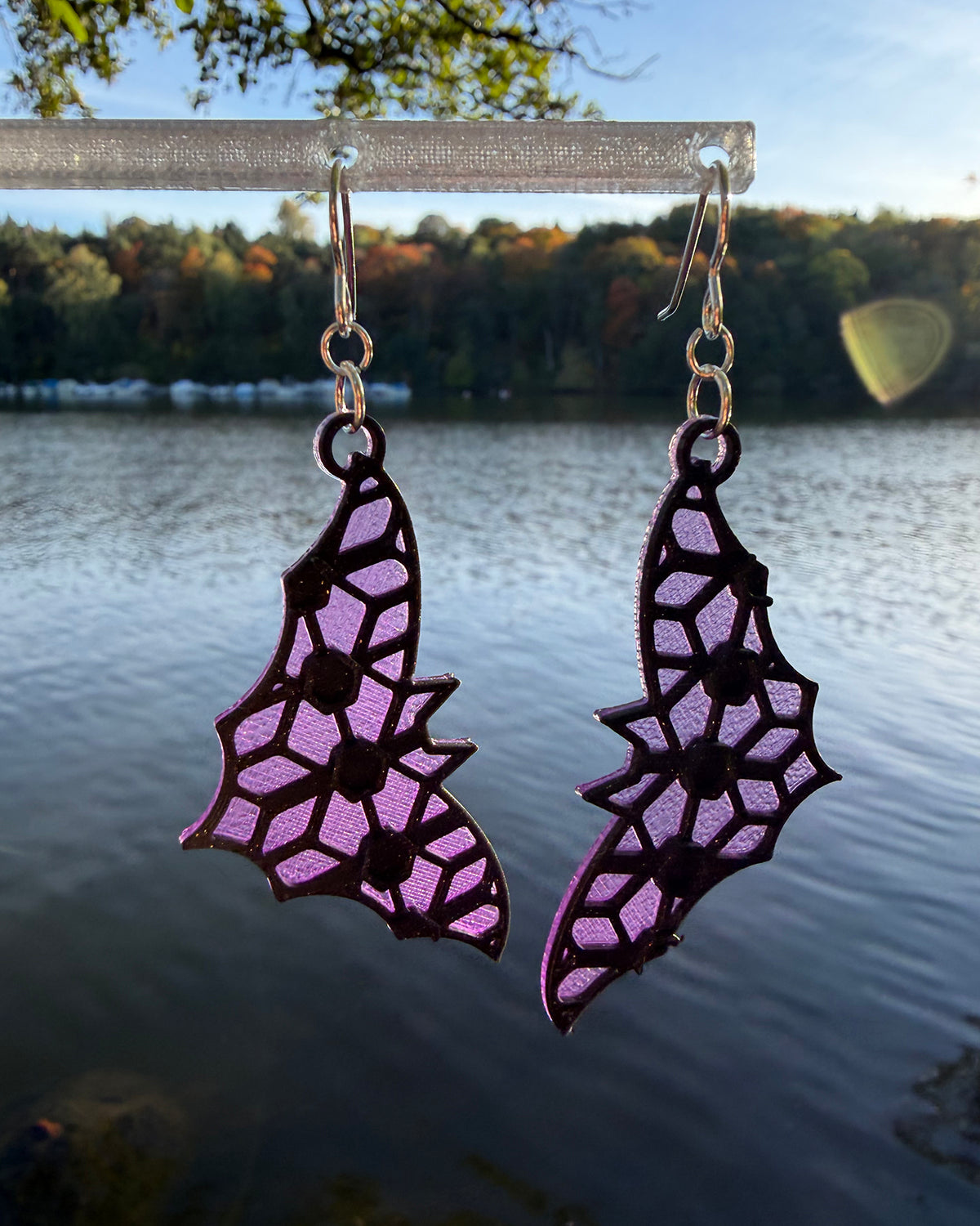 A pair of dangling earrings with a bat-shaped silhouette and a purple & black stained-glass like pattern, hanging from a clear stand against a blurred outdoor background of water and foliage.