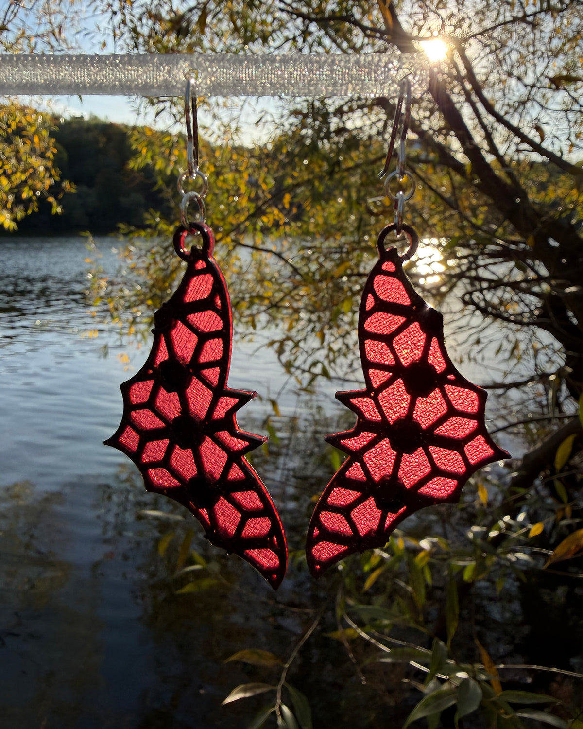 A pair of dangling earrings with a bat-shaped silhouette and a red & black stained-glass like pattern, hanging from a clear stand against a blurred outdoor background of water and foliage.