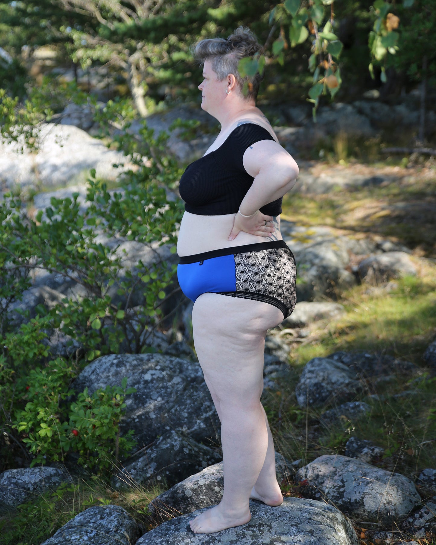 Side view of a model wearing The Mix Hipster in blue colouray. She is standing outside, with rocks and greenery around her.