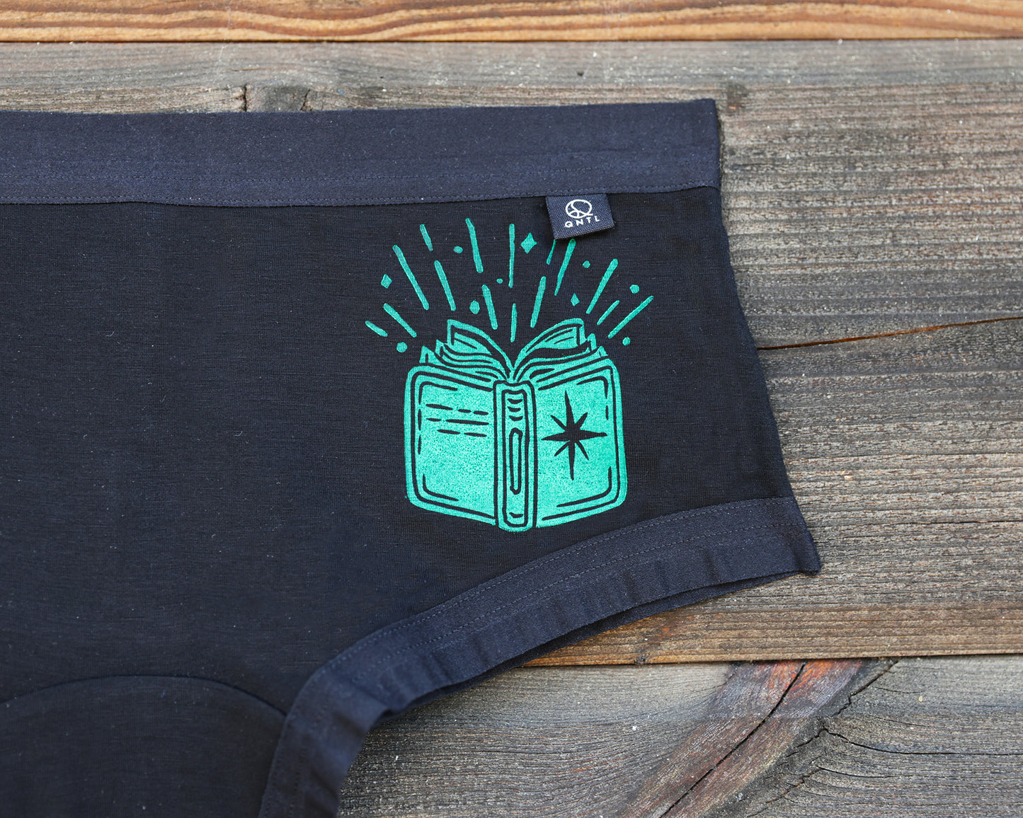 Black underwear laid flat on a rustic wooden surface, featuring a bright green graphic of an open book with radiating sparkles and a small black brand tag near the waistband.