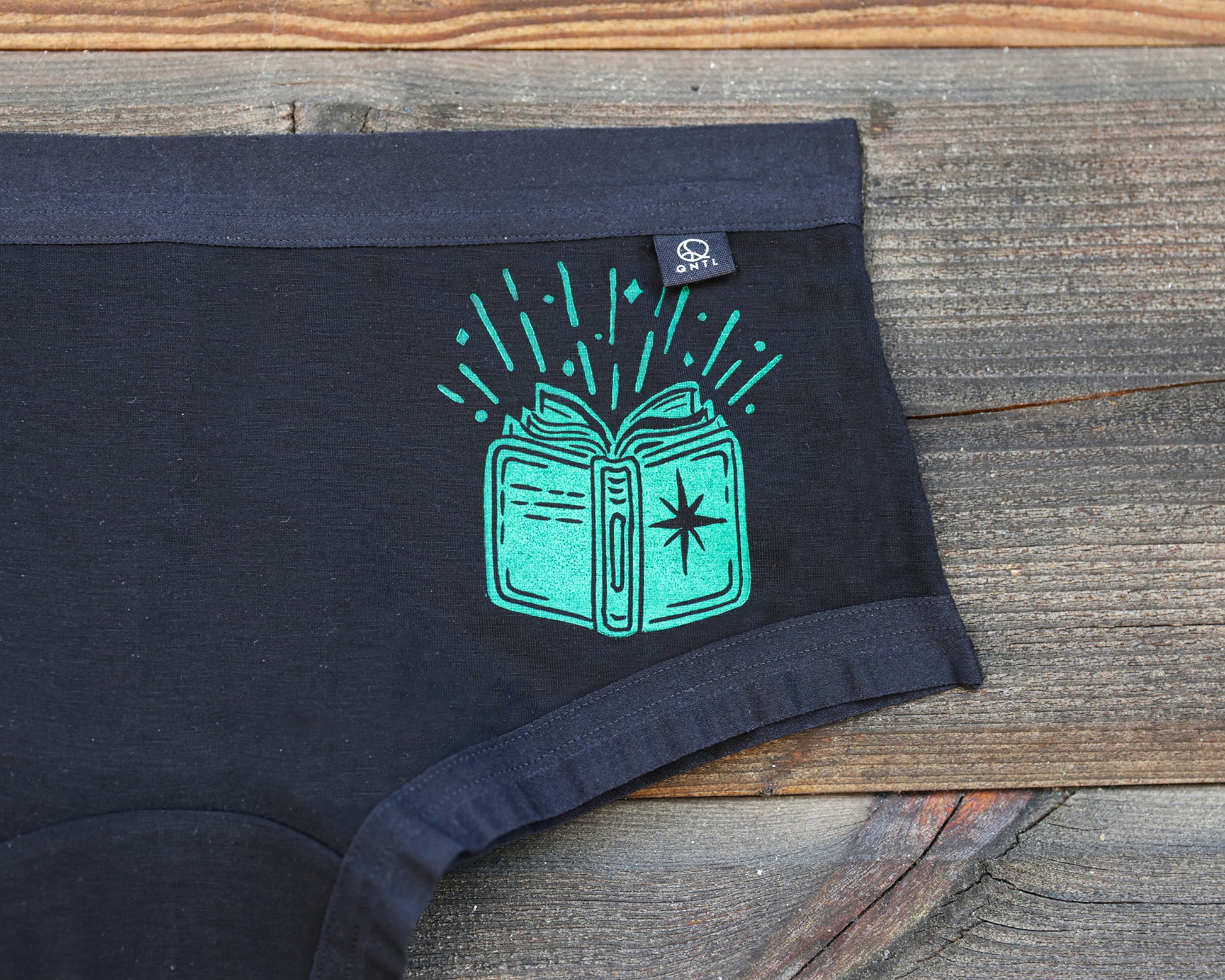 Black underwear laid flat on a rustic wooden surface, featuring a bright green graphic of an open book with radiating sparkles and a small black brand tag near the waistband.
