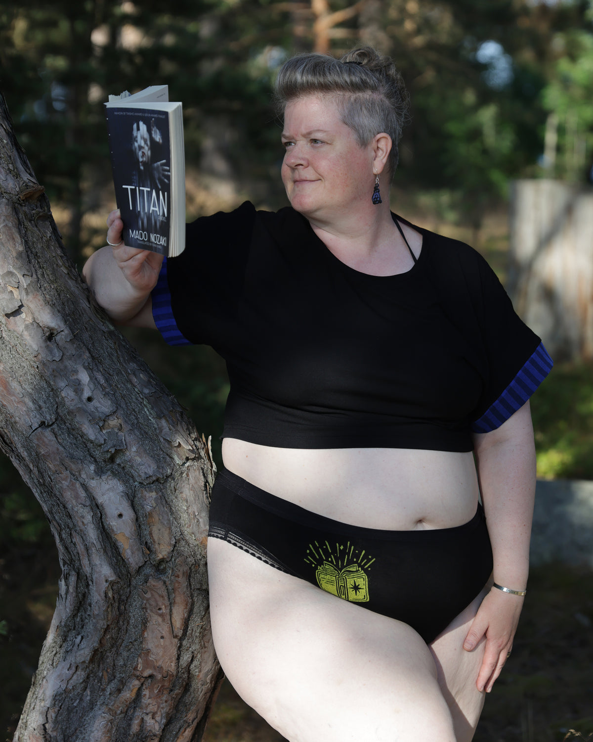 A woman leaning against a tree trunk outdoors, wearing a black cropped top with blue striped sleeves and black underwear with a yellow graphic of a book, while holding a book titled 'TITAN' by Mado Nozaki.