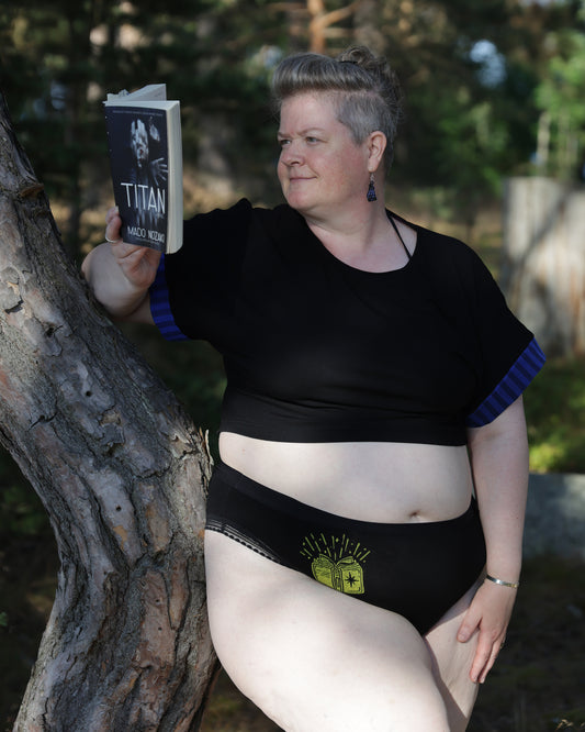 A woman leaning against a tree trunk outdoors, wearing a black cropped top with blue striped sleeves and black underwear with a yellow graphic of a book, while holding a book titled 'TITAN' by Mado Nozaki.