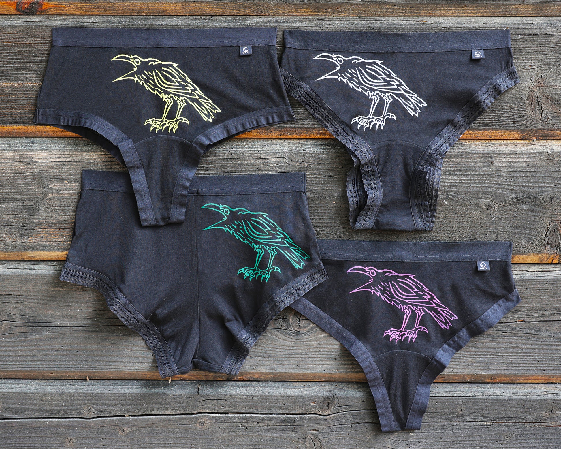 Four pairs of underwear on a rustic wooden surface, each featuring a different colored crow graphic; yellow, white, green, and magenta. This is The Crow print!
