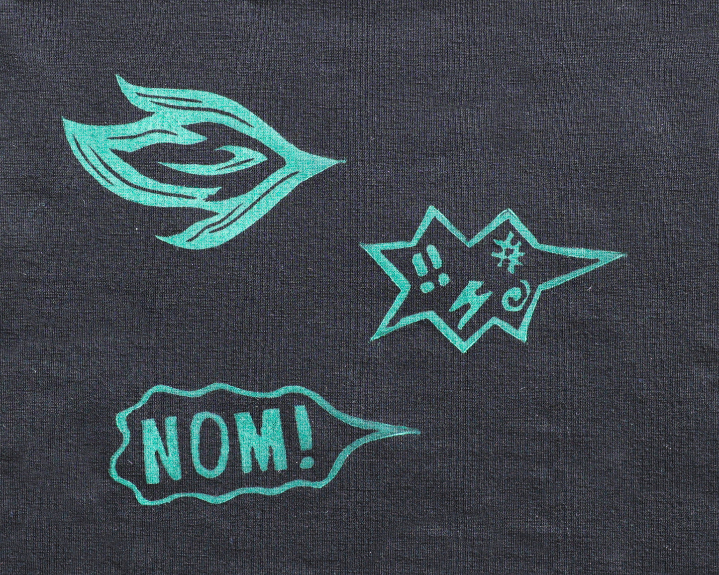 A close-up of a black jersey fabric, featuring three printed graphic elements: a stylized breath of fire, an angry speach bubble with profanities, and a speech bubble reading 'NOM!'.