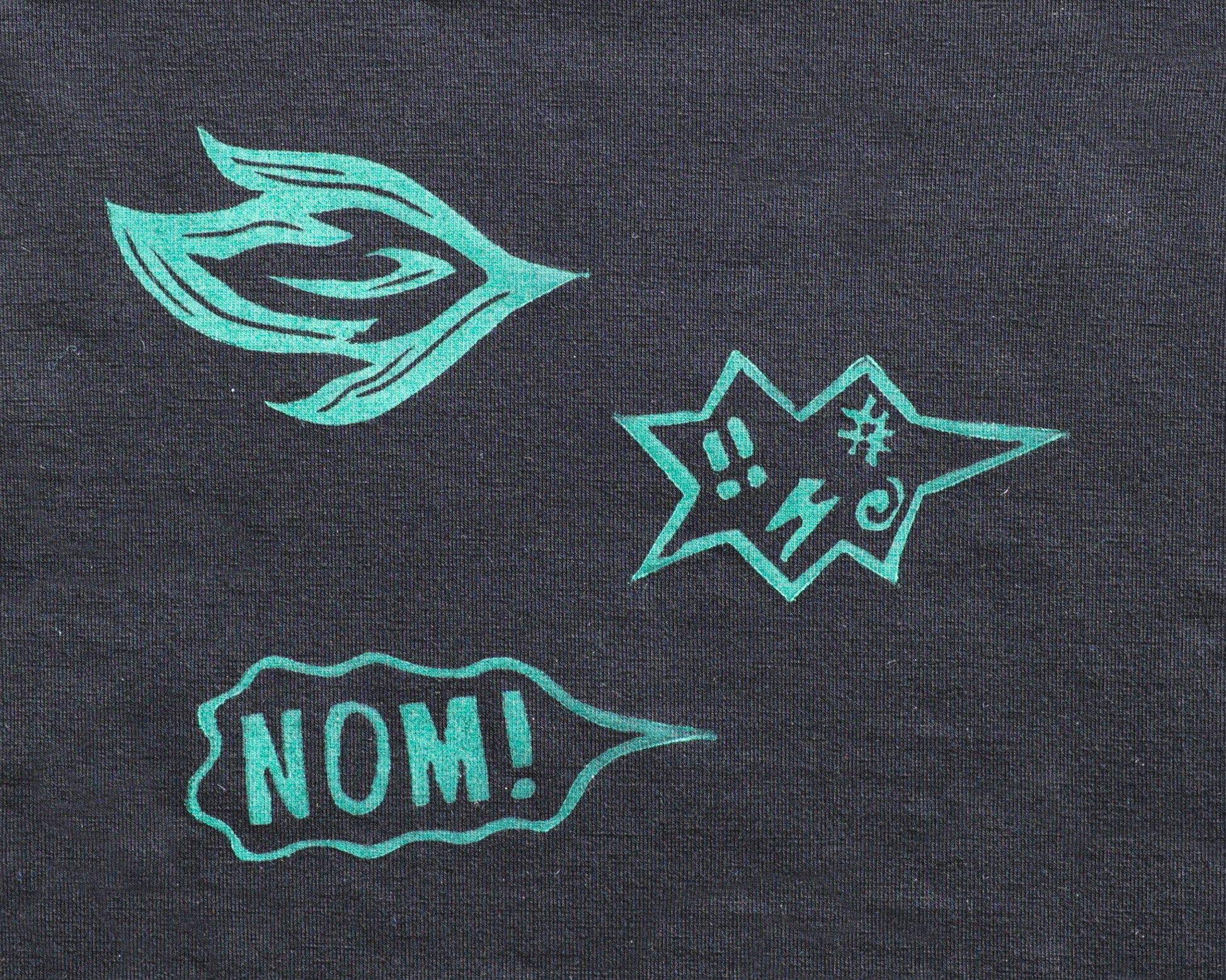 A close-up of a black jersey fabric, featuring three printed graphic elements: a stylized breath of fire, an angry speach bubble with profanities, and a speech bubble reading 'NOM!'.