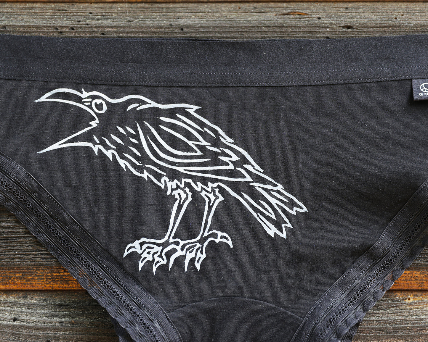 Black underwear featuring a white line art graphic of a crow on the front, laid flat on a rustic wooden surface.